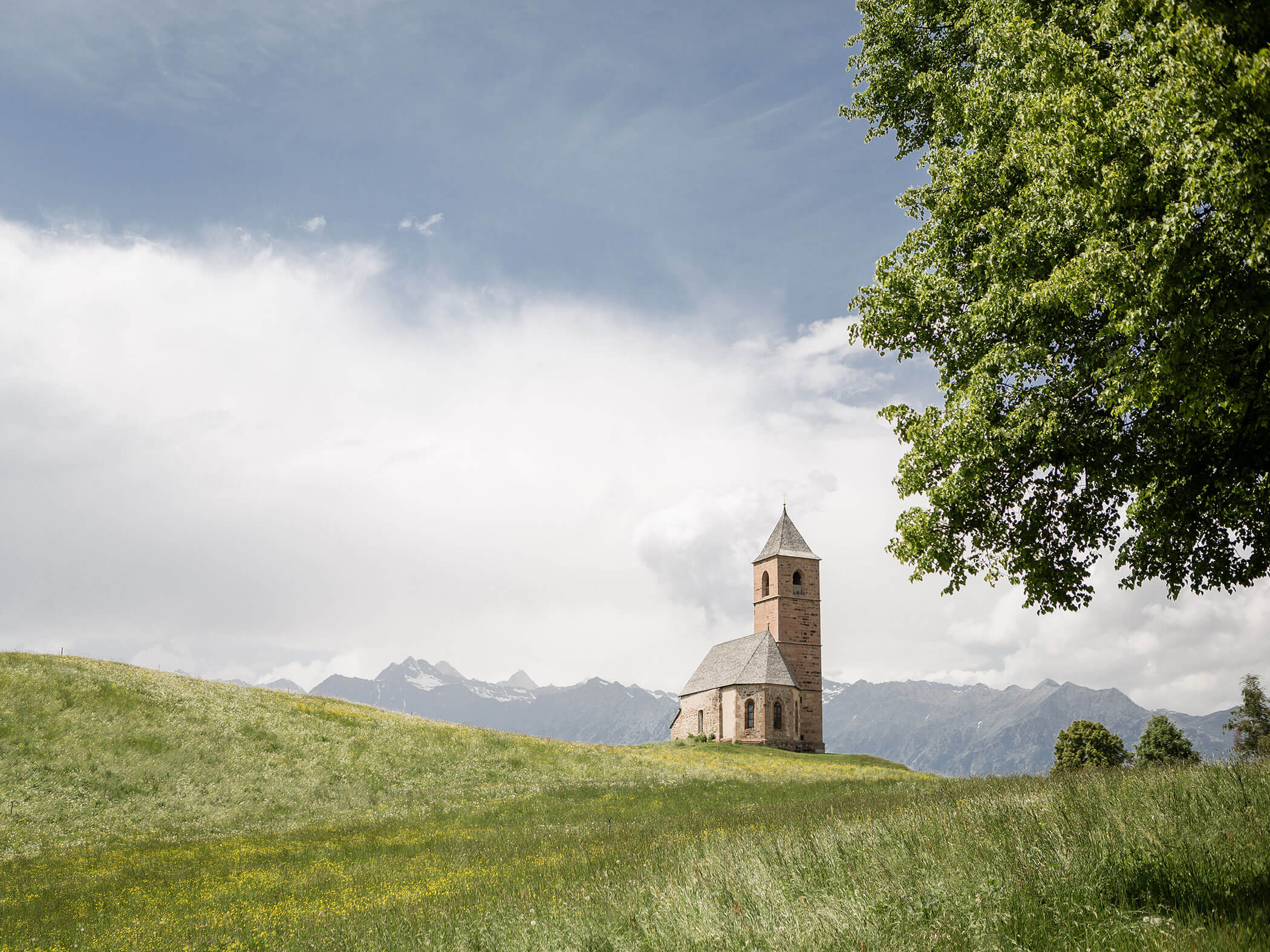 Eine kleine Kapelle steht mitten in einer Wiese, dahinter einige Berggipfel - Hotel Patriarch